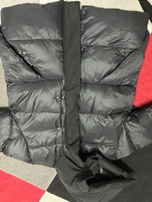 Svart Moncler pufferjacka med huva - Svart pufferjacka från Moncler med stor huva och quiltad design. Jackan har dragkedja och tryckknappar framtill, samt ett snyggt Moncler-märke på ärmen. Materialet är glansig polyester och jackan har en loose passform som ger en chill vibe.