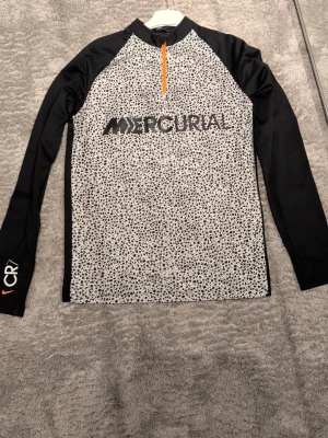 Nike Mercurial CR7 träningströja - Nike Mercurial CR7 långärmad träningströja med svartvita prickar på framsidan, svarta ärmar och orange dragkedja. Stort Mercurial-tryck på bröstet och CR7-detalj på ärmen. Perfekt för fotbollsträning.