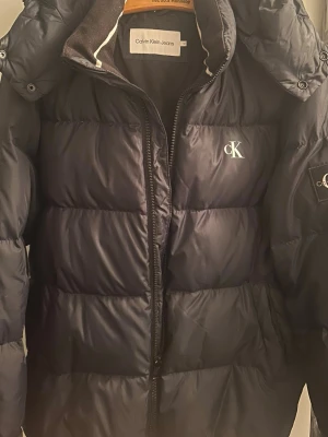Mörk blå  pufferjacka från Calvin Klein - Säljer en mörkblå  pufferjacka från Calvin Klein Jeans med hög krage och dragkedja framtill. Jackan har diskret CK-logga på bröstet och ärmen. Perfekt för kalla dagar och riktigt snygg till streetwear-stilen. 