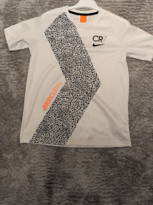 Nike CR7 Mercurial t-shirt vit - Snygg vit Nike t-shirt med CR7- och Mercurial-tryck. T-shirten har korta ärmar och ett svart prickigt mönster diagonalt över framsidan samt orange detalj. Perfekt för dig som gillar sportig stil och Cristiano Ronaldo.