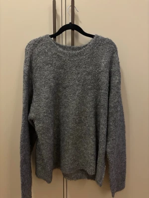 Grå stickad tröja från Woodbird - Mysig grå stickad tröja från Woodbird med rund hals och lång ärm. Tröjan har en fluffig, lite luddig textur som ger en avslappnad vibe och passar perfekt till lager på lager. Enkel och stilren design som är lätt att matcha.