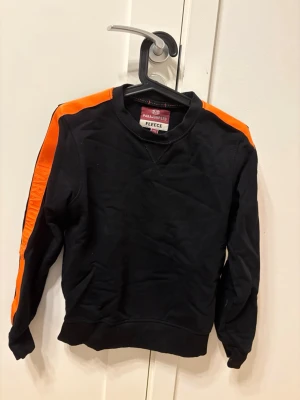 Svart fleece tröja Parajumpers Y-S - Cool svart fleece tröja från Parajumpers med orangea detaljer längs ärmarna och ett stort broderat PJS-märke på ena armen. Tröjan har rund hals och långa ärmar, perfekt för dig som gillar sportig och street-inspirerad stil.