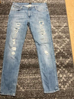 Ljusblå slitna Lee jeans - Säljer ett par ljusblå jeans från Lee med snygga slitningar och hål på både fram- och baksida. Klassisk femficksmodell med Lee-lapp bak i brunt. Jeansen har rak passform och är tillverkade i mjukt bomullsdenim.