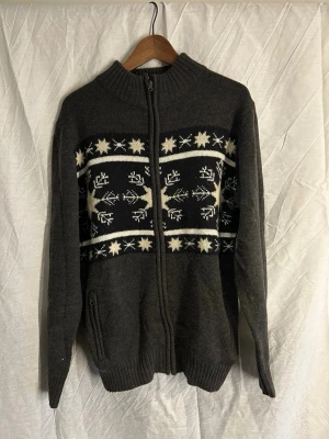 *Unik* stickad cardigan  - Mysig mörkgrå kofta med dragkedja och ribbad krage. Framtill finns ett klassiskt vintermönster i vitt med renar och snöflingor. Koftan är stickad och har långa ärmar samt fickor på sidorna. Perfekt för kalla dagar och vintermys.