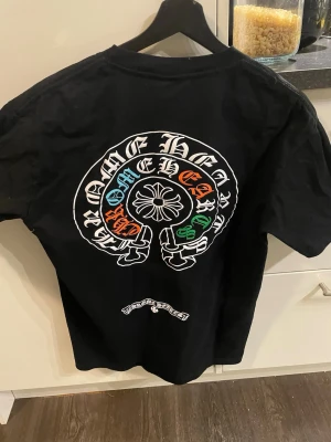 Svart Chrome Hearts t-shirt - Säljer en svart t-shirt från Chrome Hearts med stort tryck i vitt, blått, orange och grönt på ryggen samt mindre logga på bröstet. Klassisk rund hals och korta ärmar. Tillverkad i mjuk bomull för skön passform.
