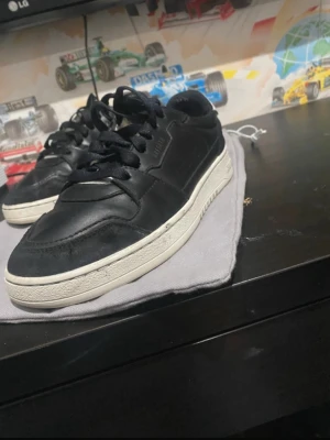 Svarta sneakers från Axel Arigato - Säljer ett par svarta sneakers från Axel Arigato med vit sula och klassisk låg profil. Skorna har snörning och är tillverkade i skinn med diskret logga på sidan. Perfekta för dig som gillar stilrena och tidlösa sneakers.