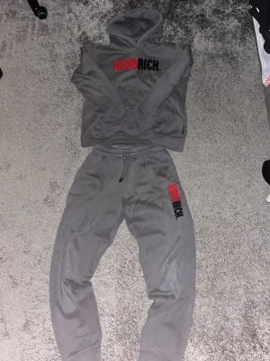 Grå mjukisbyxor från Hoodrich  - Gråa mjukisbyxor från Hoodrich med broderad logga i rött och svart på ena benet. Byxorna har resår och snörning i midjan samt muddar vid bensluten. Sköna och stilrena, perfekta för chill eller streetwear.