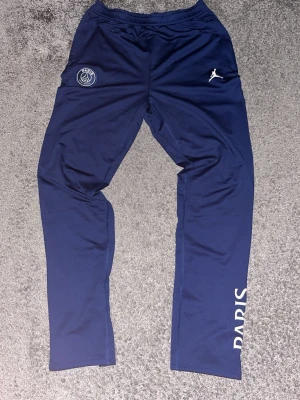 PSG x Jordan träningsbyxor marinblå - Marinblå träningsbyxor från PSG x Jordan med vita detaljer och tryckt Paris-text på benet. Byxorna har elastisk midja, dragkedja vid benslut och är tillverkade i ett mjukt, syntetiskt material. Perfekta för fotbollsträning eller chill.