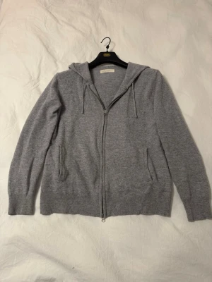 Grå cashmere hoodie från Sayless - Mysig grå hoodie från Sayless i mjuk cashmere. Tröjan har dragkedja framtill, två fickor och huva med snörning. Perfekt för dig som gillar stilrena och bekväma plagg med en lyxig känsla.