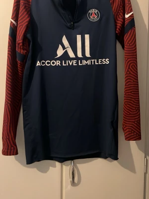 PSG Nike Dri-Fit träningströja S - Säljer en PSG träningströja från Nike i Dri-Fit material. Tröjan är marinblå med röda, mönstrade ärmar och har Paris Saint-Germain-logga på bröstet. Halv dragkedja framtill och vit text med 'Accor Live Limitless' på magen. Perfekt för fotbollsträning.