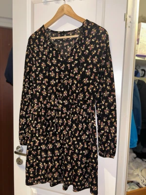 Blommig svart kortklänning Gina Tricot - Supersöt svart kortklänning från Gina Tricot med små blommor i gult, vitt och rosa. Klänningen har lång ärm, v-ringning och lätt rynkad midja för en avslappnad siluett. Perfekt för dig som gillar en feminin och chill vibe.