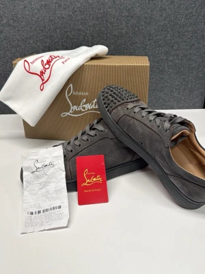 Grå mocka sneakers från Louboutin - Säljer ett par grå sneakers från Christian Louboutin i mocka med coola nitar på tån och klassisk röd sula. Skorna har snörning och en låg profil, samt beige innersula. Perfekt för dig som vill sticka ut med exklusiva detaljer. Nypris ca 10k. Skriv för frågor och självklart så är de äkta!