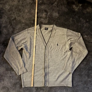 Grå kofta från Polo Ralph Lauren M - Klassisk grå kofta från Polo Ralph Lauren med v-ringning och knappar framtill. Liten broderad logga på bröstet. Tillverkad i mjukt material, perfekt för lager-på-lager. Långärmad och stilren design som funkar året runt.