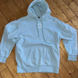 Ljusblå Overtime hoodie med tryck - Ljusblå hoodie från Overtime med vit logga på bröstet och texten OVERTIME tryckt längs ena ärmen. Klassisk känguruficka framtill, huva med dragsko och mjukt material som känns skönt mot huden. Perfekt för chill dagar eller när du vill ha en clean streetstyle.