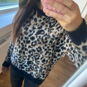 Leopardmönstrad stickad tröja - Säljer en stickad tröja med leopardmönster i svart, beige och bruna toner. Tröjan har rund halsringning och svarta ribbade muddar vid ärmslut och nederkant. Perfekt för dig som gillar djurmönster och vill ha en mysig, snygg look.