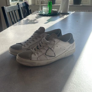 Philippe Model skor - Snygga vita sneakers från Philippe Model med grå mockadetaljer på tån och svart hälparti. Skorna har snörning, rund tå och en platt sula. Tillverkade i skinn och mocka, med en ikonisk sköld på sidan. Perfekta för dig som gillar stilrena och trendiga sneakers. Skorna är i stl 46