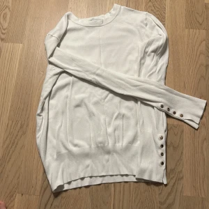 Vit långärmad tröja med knappar - Vit långärmad tröja med rund hals och dekorativa guldfärgade knappar vid ärmslut och nederkant. Mjuk och tunn stickad känsla, perfekt för lager-på-lager. Enkel och stilren design som passar till det mesta.