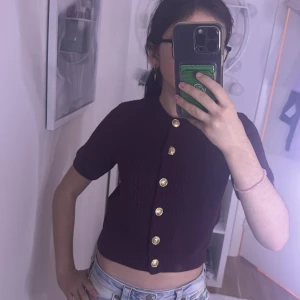 Vinröd stickad blus från H&M - Snygg vinröd stickad blus från H&M med korta ärmar och croppad passform. Framsidan har sex guldiga knappar som ger en cool touch. Materialet är stickat och känns mjukt mot huden. Perfekt för dig som gillar detaljer och vill ha en unik topp i garderoben.