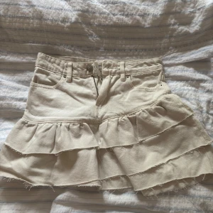 Beige volangkjol i jeans från Perfect Jeans - Supersöt beige kortkjol i jeansmaterial från Perfect Jeans. Kjolen har flera volanger, råa kanter och klassiska jeansdetaljer som fickor och bälteshällor. Perfekt för dig som gillar en trendig och avslappnad stil.