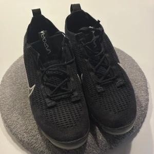 Nike Vapormax Flyknit svarta sneakers - Svarta Nike Vapormax Flyknit sneakers med vit swoosh på sidan och stickad ovandel. Skorna har snörning, genomskinlig sula och dragflik bak. Perfekta för dig som gillar sportig och modern stil.