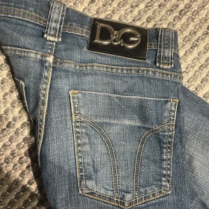 Blå jeans från Dolce & Gabbana D&G - Snygga blå jeans från Dolce & Gabbana med klassisk D&G-logga på bakfickan och läderpatch i midjan. Jeansen har raka ben, kontrastsömmar och femficksdesign. Perfekt för dig som gillar stilrena och ikoniska denimplagg. Industriell hål vid ena knäet