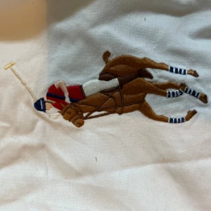 Vit skjorta från Ralph Lauren - Vit skjorta från Ralph Lauren med stor broderad polospelare på bröstet och en mörkblå siffra 3 på ena ärmen. Klassisk krage, knäppning framtill och lång ärm. Skjortan är i bomull och har en clean och sportig look.