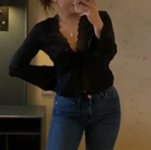 Svart omlottblus med spets från Nelly - Svart omlottblus från Nelly med långa ärmar, djup v-ringning och fina spetsdetaljer längs kanten. Blusen knyts framtill med två smala band och har en lätt glansig yta💕