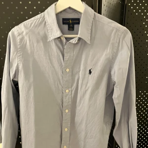 Ljusblå skjorta från Ralph Lauren - Klassisk ljusblå skjorta från Ralph Lauren med broderad marinblå logga på bröstet. Skjortan har lång ärm, knappar framtill och en snygg custom fit-passform. Perfekt för dig som gillar stilrena och tidlösa plagg. Skjortan är ljusblå, sista bilden visar färgen bäst! Storlek 18 motsvarar ca 163-168cm. Nypris ca 1190,- 