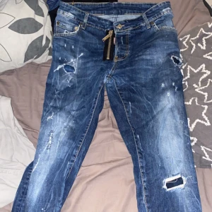Dsquared2 blå  - Blå jeans från Dsquared2 med coola slitningar, hål och färgstänk. Snygg, stor dragkedja framtill och klassiska kontrastsömmar. Jeansen har en modern, streetig look och är tillverkade i denim med detaljer som sticker ut.
