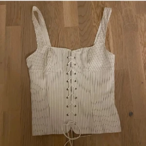 Vit randig korsett med snörning - Supersnygg vit korsett med tunna svarta ränder och snörning framtill. Toppen är använd fåtal gånger och ser som ny ut. Sytt upp banden en halv cm på varsin sida då banden är rätt så långa, går att sprätta upp jätte enkelt!