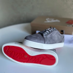 Grå mockasneakers från Christian Louboutin - Oanvända grå sneakers från Christian Louboutin i mjuk mocka med klassisk röd sula och vita kanter. Skorna har rund tå, snörning och beige innersida i skinn. Perfekta för dig som vill ha en stilren och lyxig look med en ikonisk touch.
