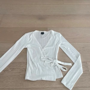 Vit omlottopp från Gina - Supersöt vit omlottopp från Gina Tricot med långa ärmar, knyt i midjan och fina spetsdetaljer längs v-ringningen. Toppen är i ett fint mönster. Den är iprincip oanvänd så i väldigt bra skick