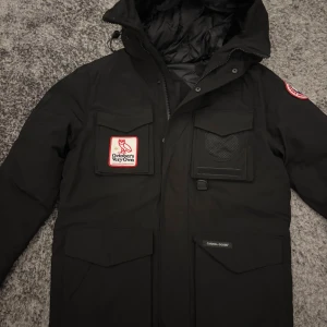 Svart dunjacka Canada Goose x OVO - Svart dunjacka från Canada Goose i samarbete med October's Very Own. Jackan har flera fickor framtill, patchar på bröstet och ärmen, samt huva. Fodrad insida och klassisk Canada Goose-detalj på ärmen. Perfekt för kalla dagar.