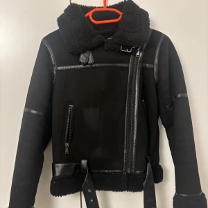Svart teddyjacka från Zara med bälte - Säljer en svart teddyjacka från Zara med mjuk fuskpäls på insidan och kragen. Jackan har detaljer i skinnimitation, sned dragkedja, bälte i midjan och två fickor med dragkedja. Perfekt för kalla dagar och riktigt snygg till streetwear.