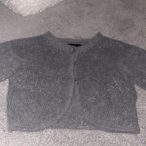 Grå stickad kofta från Vero Moda - Mysig grå stickad kofta från Vero Moda i kort modell. Koftan har rund halsringning, en knapp framtill och är stickad i ett mjukt material. Perfekt att slänga över en topp för extra värme under kyliga dagar.