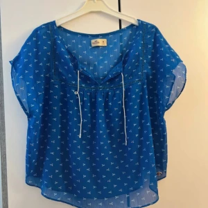 Blå blus med småmönster från Hollister - Söt blå blus från Hollister med små vita och rosa blommönster. Blusen är luftig och har korta ärmar, v-ringning med knytband och transparent lätt material. Perfekt för varma dagar och har en liten broderad Hollister-logga nertill.