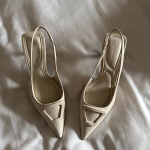 Beige pumps med spetsig tå  - Snygga beige pumps från Primark med spetsig tå och låg klack. Skorna har en öppen häl med smal rem och dekorativ detalj framtill. Tillverkade i glansigt syntetmaterial och har en elegant, modern form som sticker ut.