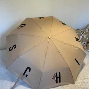 New Moschino beige umbrella - Unused Moschino beige umbrella. Original price 95€.  • Product number: ZZO281S18-B00  • Handle material: plastic • Outer fabric material: 100% polyester  • Length: 48 cm • Diameter: 99 cm 