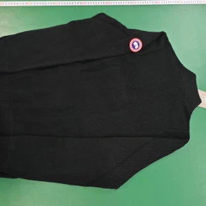 Svart quiltad dunväst från Canada Goose - Svart dunväst från Canada Goose med quiltad framsida och stickade sidor. Västen har hög krage, dragkedja och klassisk logotyp på axeln. Perfekt för lager-på-lager och ger en clean, sportig look.
