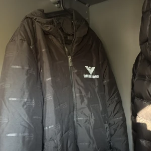 Svart jacka  från Emporio Armani - Svart tjock jacka  från Emporio Armani med huva och dragkedja framtill. Jackan har logotypen tryckt på bröstet och huvan, samt diskreta mönster i tyget. Snygg och sportig look, perfekt för dig som gillar stilrena märkesplagg. Den har ett hål i en av fickorna men enkelt att fixa.