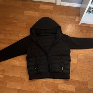Svart cardigan med huva - Svart pufferjacka med huva och dragkedja framtill. Jackan har quiltad framsida och rygg, släta ärmar och två fickor med dragkedja. Perfekt för kyligare dagar och har en clean, stilren look. Jackan är M men passar lika bra till S.