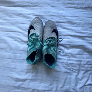 Nike Phantom Elite fotbollsskor - Säljer ett par Nike Phantom GT2 Elite fotbollsskor i grått och turkos med svarta och lila detaljer. Skorna har snörning, högre strumpkrage och plös, samt dobbar i lila och svart för bästa grepp på planen. Ovandelen är i syntetmaterial för bra bollkontroll. Storlek 42,5