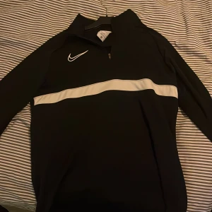 Nike svart Dri-Fit half zip tröja - Svart långärmad Nike Dri-Fit tröja med vit rand över bröstet och klassisk Swoosh-logga. Perfekt till mjukisbyxor