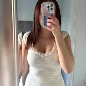 Vit figurnära topp med spetsdetalj - Säljer en vit, figurnära topp med korta ärmar och söt spetskant vid urringningen. Toppen har en liten rosett framtill och lätt rynkning vid bysten. Perfekt för dig som gillar en feminin och enkel stil.