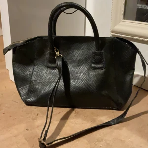 Svart axelväska i skinnimitation - Snygg svart axelväska med strukturmönster och guldfärgade detaljer. Väskan har både handtag och justerbar axelrem, perfekt för att bära på olika sätt. Stängs med dragkedja och rymmer allt du behöver till vardagen.