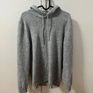 Grå stickad hoodie med huva - Mysig grå stickad hoodie med huva och snörning. Tröjan har ribbade muddar vid ärmslut och nederkant.