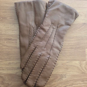 Bruna skinnhandskar med dekorativa sömmar - Snygga bruna handskar i skinn med dekorativa, synliga sömmar längs fingrarna och runt kanterna. Handskarna har en klassisk form och är perfekta för kyligare dagar. De är mjuka och följsamma, vilket gör dem bekväma att bära. Fodrade med ull/bomull. Nyskick, aldrig använda. 