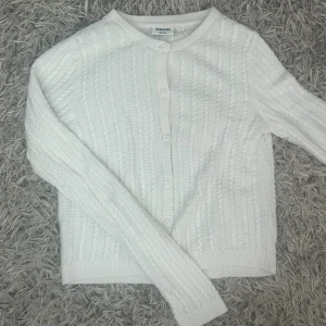 Vit kabelstickad kofta från Kappahl - Supersöt vit kabelstickad kofta från Kappahl med rund halsringning och knappar framtill. Långärmad modell med ribbade muddar och nederkant. Perfekt att slänga över en topp när det blir lite kyligt. Stl 158/164/xxs