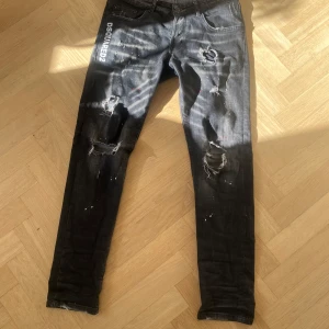 Svarta Dsquared2 jeans äkta! - Säljer ett par svarta jeans från Dsquared2 med tydliga slitningar och hål på benen, samt vit Dsquared2-logga på framsidan och röd ICON-detalj bak. Jeansen har smal passform och är tillverkade i bomull. Perfekt för dig som gillar en edgy streetstil.