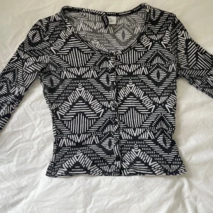 Svartvit mönstrad blus med knappar - Säljer en svartvit blus från Gina Tricot med grafiskt mönster och rund halsringning. Toppen har långa ärmar, knappar framtill och är figurnära i modellen. Perfekt för dig som gillar unika prints och vill sticka ut lite extra.